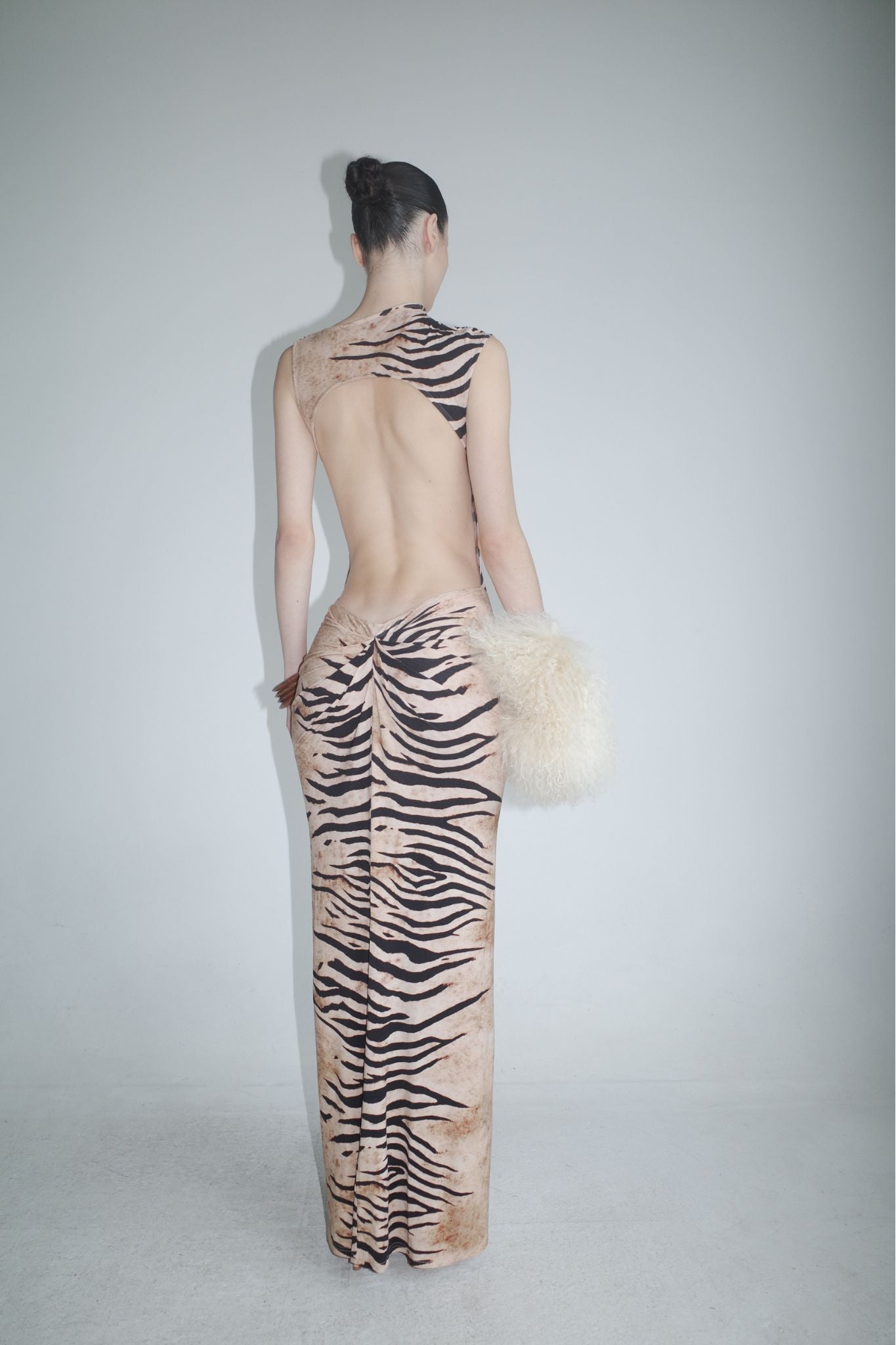 A person faces away, wearing the I.AM.GIA ELIANA MAXI DRESS - WILD SIDE, a floor-length backless animal print dress. Their hair is in a bun and they hold a large fluffy cream handbag, standing before a plain light wall.
