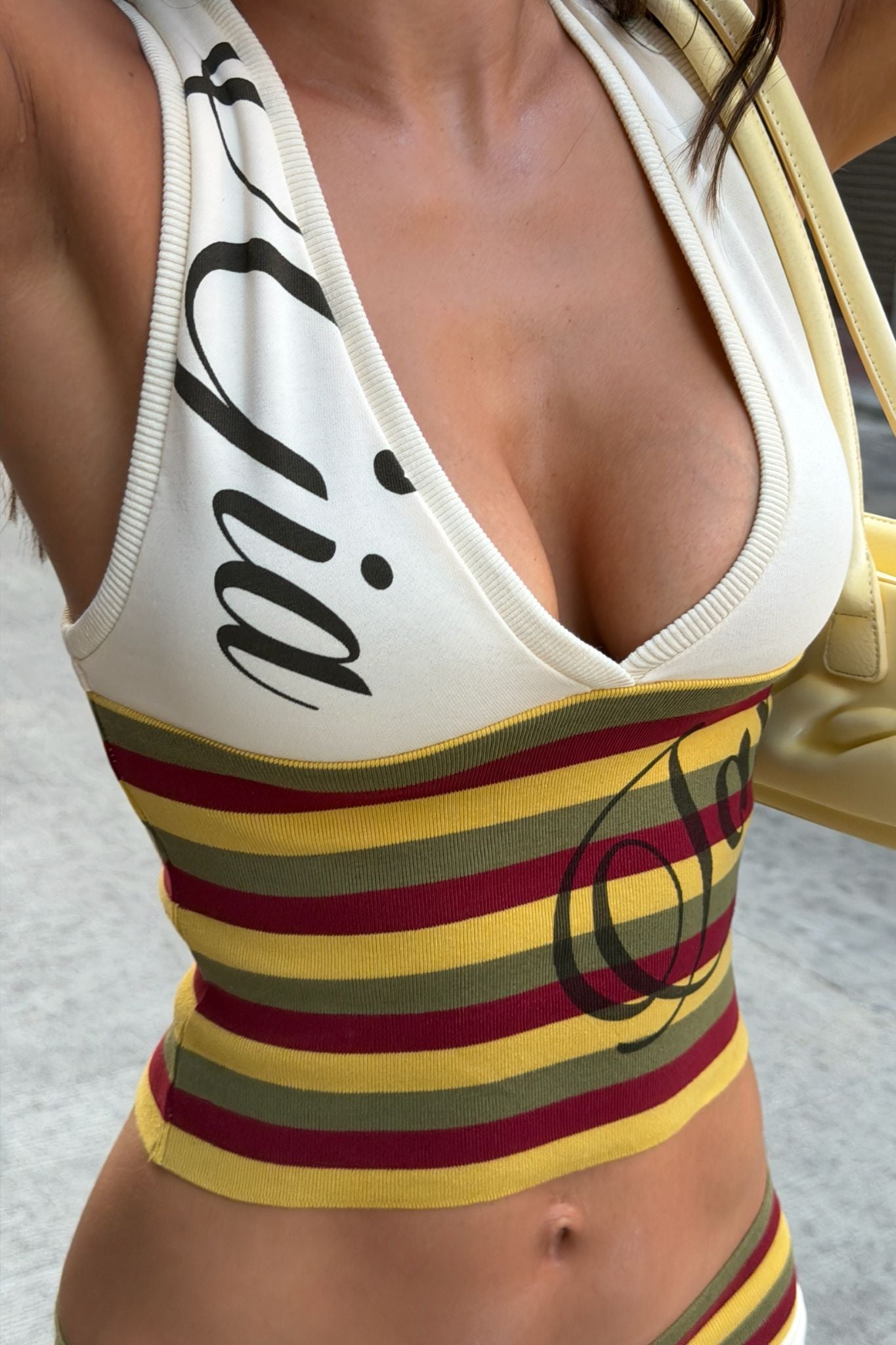 A person wearing the I.AM.GIA CANDI TOP - CREAM, a sleeveless V-neck racerback with yellow, red, and dark green stripes plus black text, holds a yellow handbag outdoors. The photo is cropped at the shoulders.
