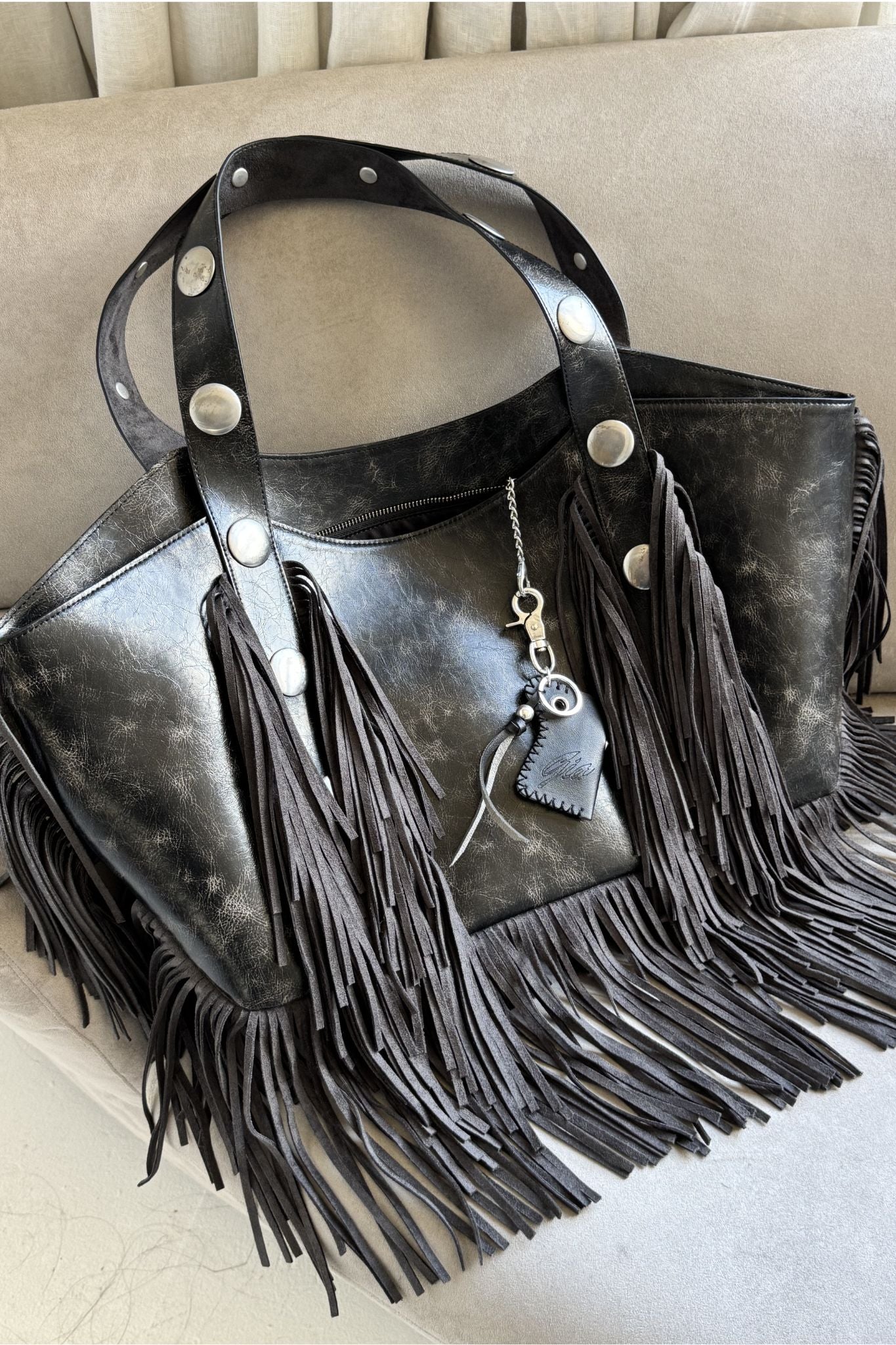 The CHROMA FRINGE BAG - BLACK by I.AM.GIA features long faux leather fringes on the sides and bottom, large silver studs on the handles, and a keychain with a small black charm, all set against a light fabric background.
