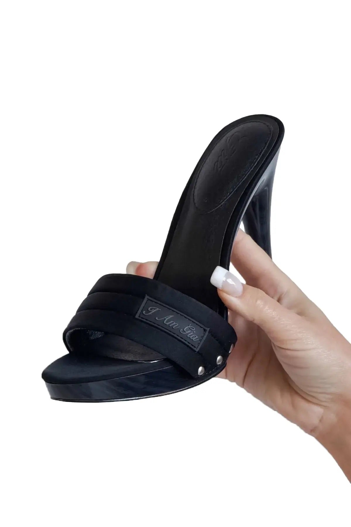 A hand with white-tipped nails holds the I.AM.GIA NICKY HEEL in black neoprene, a stiletto sandal with an open toe, wide padded vamp strap, metallic logo plate, and side studs.