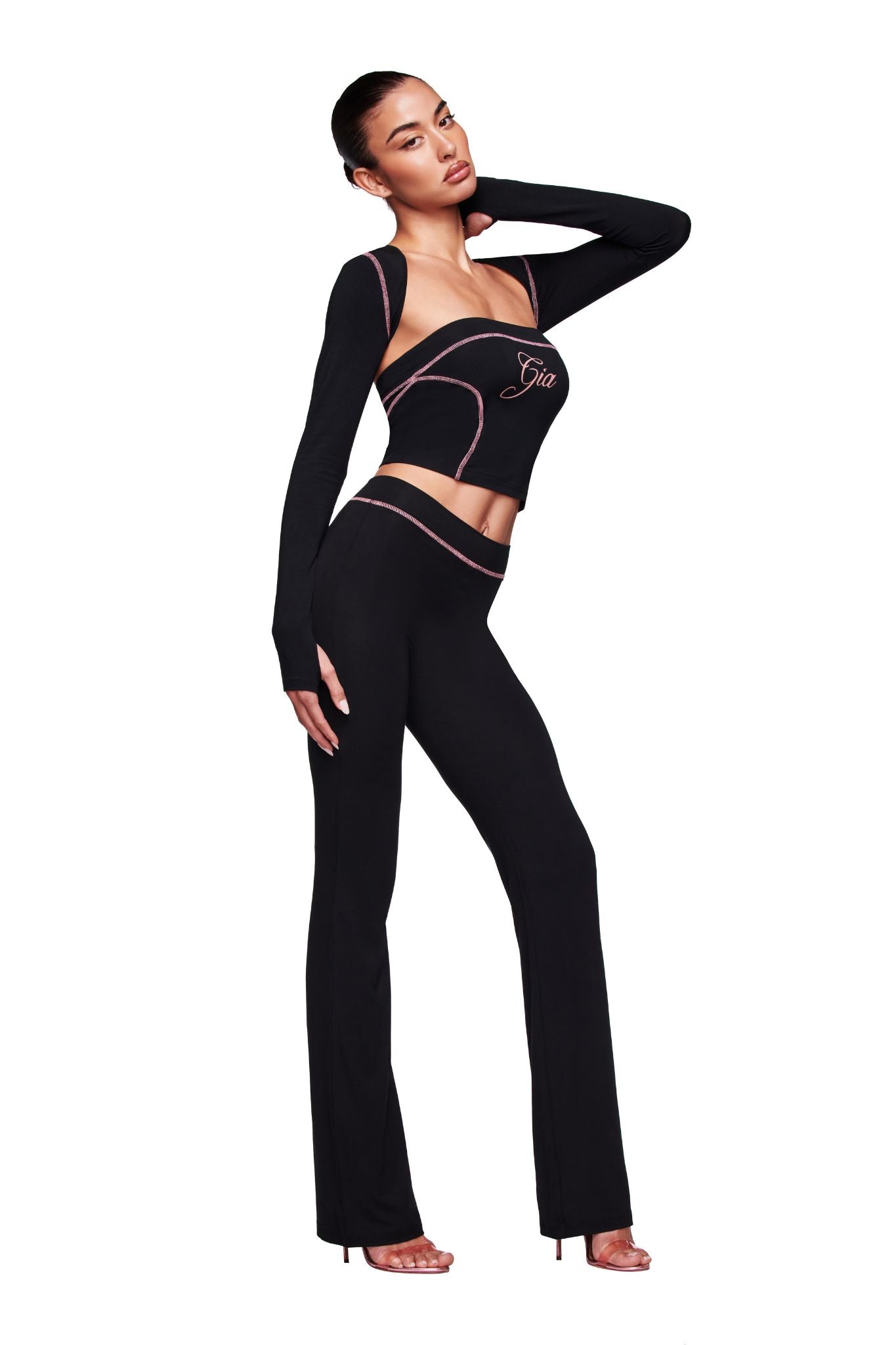 A woman models the I.AM.GIA BLARE SHRUG in black, layered over a fitted two-piece with pink trim and “Gia” on the top. She poses confidently, one hand on her neck and the other on her hip, against a plain white backdrop.