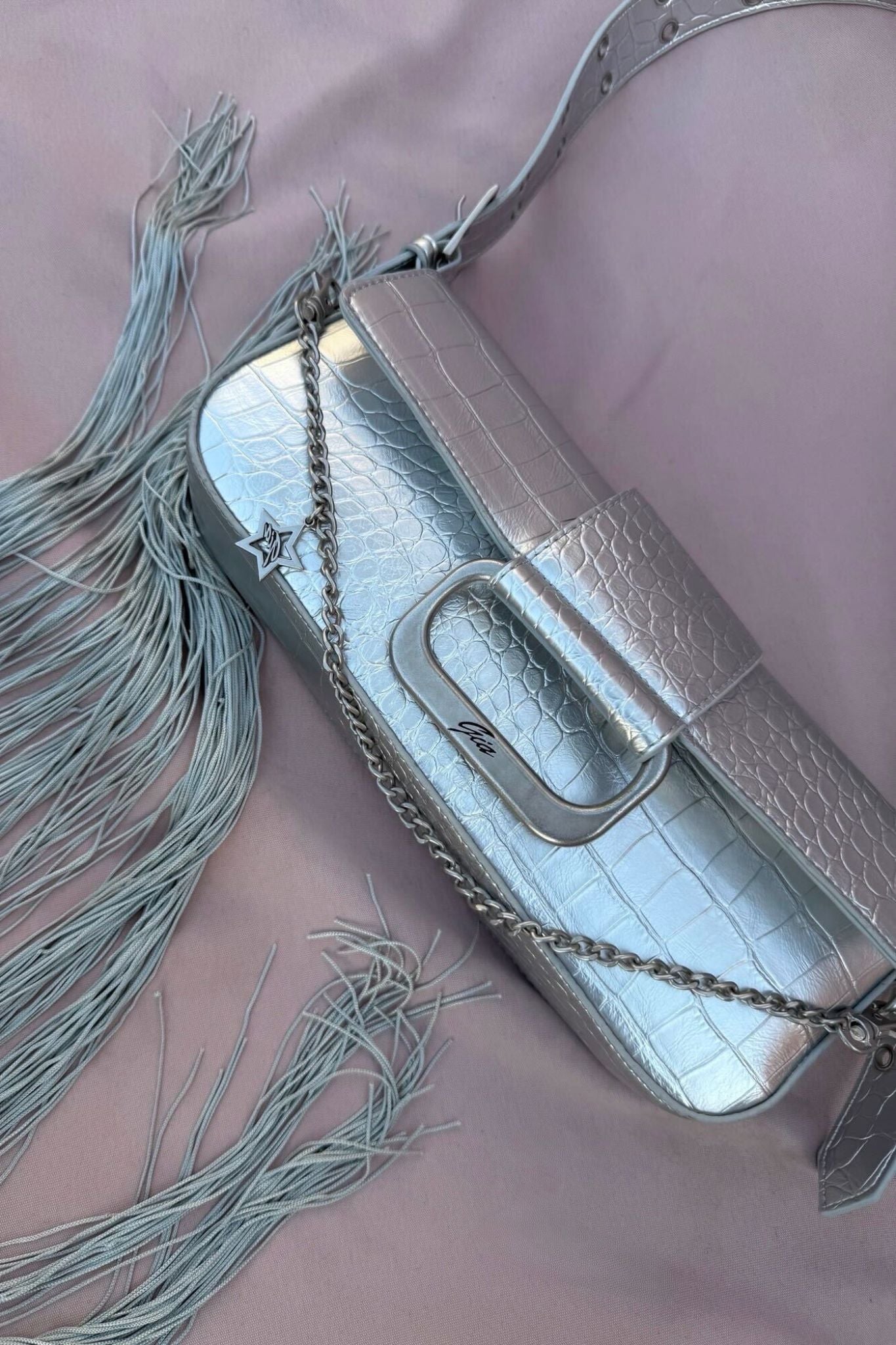 The MILA METALLIC BAG - SILVER METALLIC by I.AM.GIA, a silver mini shoulder purse with faux crocodile texture, chain strap, and long light grey fringe, is shown on a soft pink background with subtle I.AM.GIA branding.