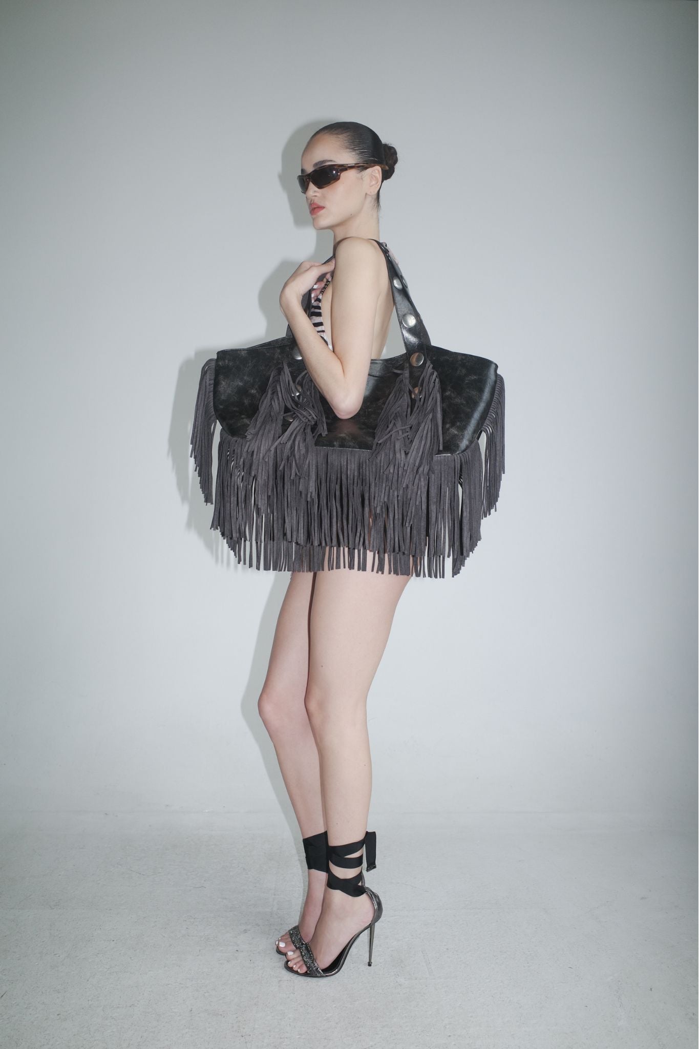A woman in high heels and sunglasses stands in profile with a sleek bun, posing against a plain background while carrying the I.AM.GIA CHROMA FRINGE BAG - BLACK, an oversized faux leather tote with distressed black fringe.