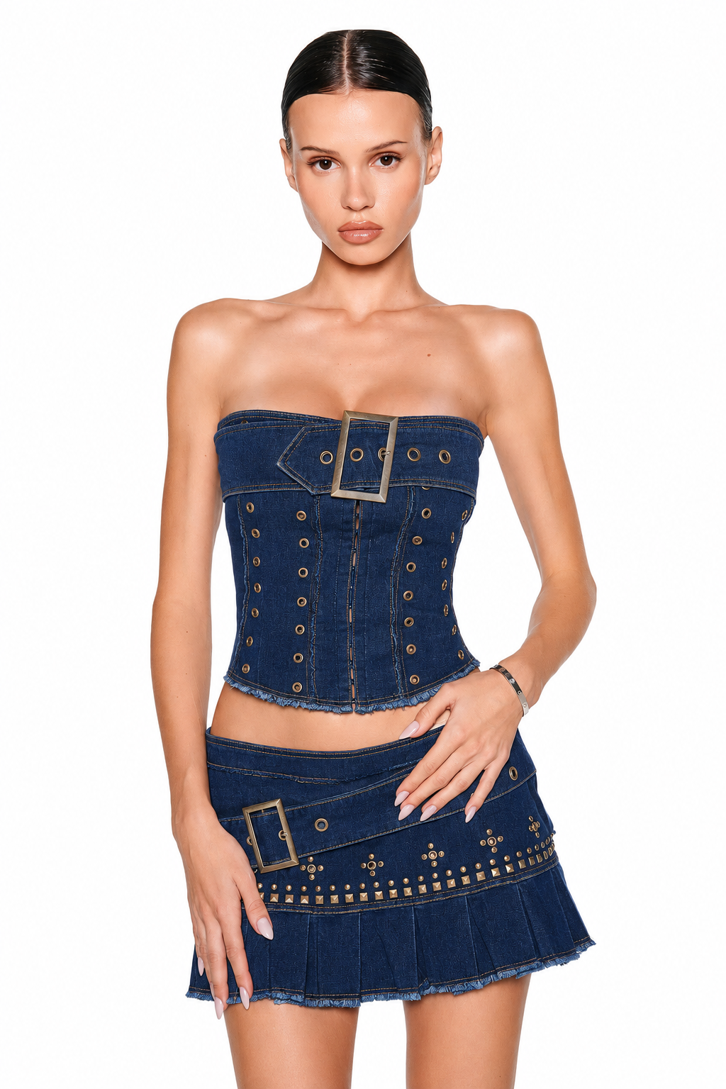 A woman poses against a white background wearing a strapless denim corset top with metallic grommets and the MASON MINI SKIRT in dark denim by I.AM.GIA, which features studded details and a large buckle.