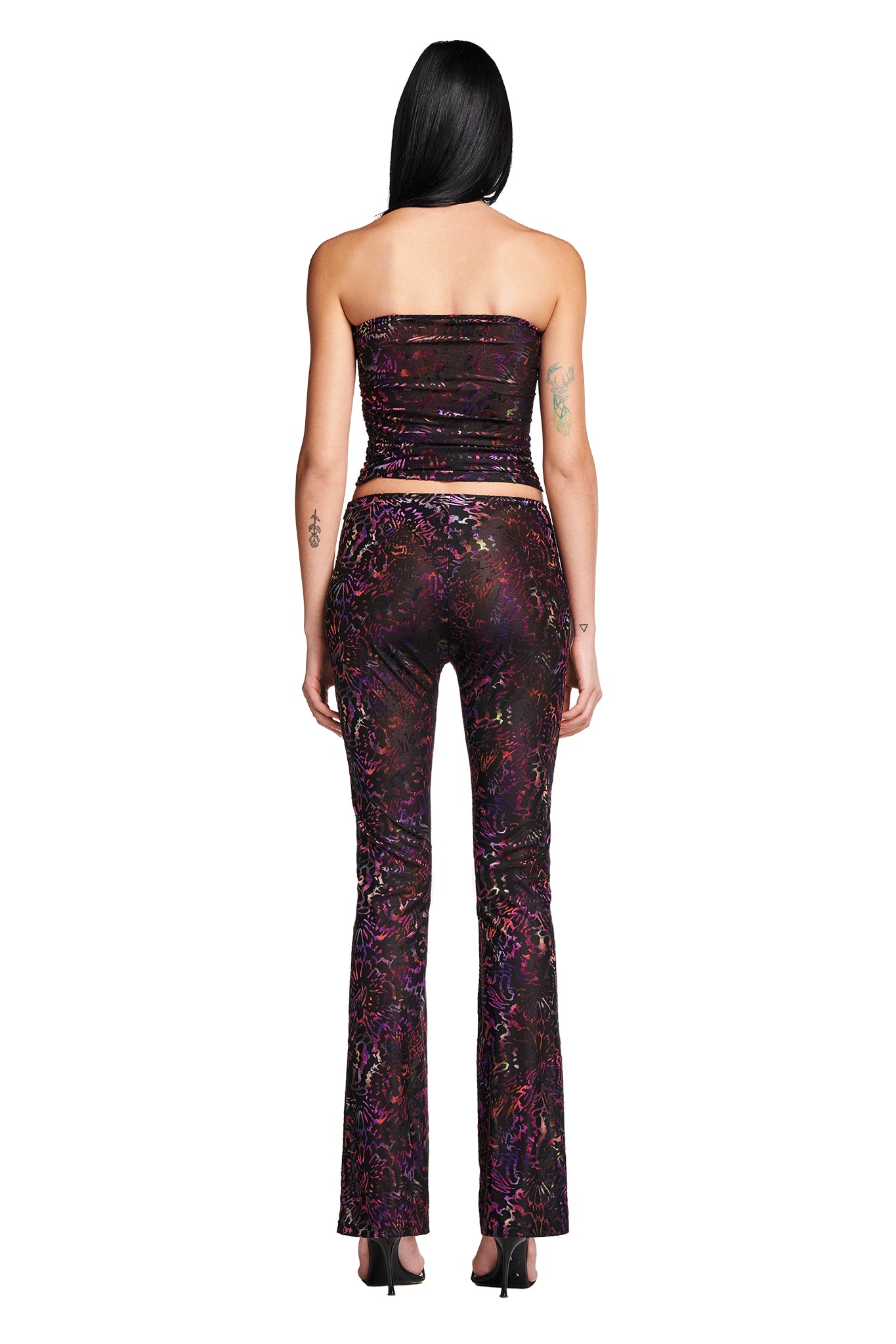 A woman with long dark hair stands facing away from the camera, wearing a stylish I.AM.GIA two-piece outfit. She has on a strapless crop top and the high-waisted, flared AMINA PANT - ANIMAL BURN OUT. The ensemble features a colorful and intricate abstract pattern on a dark background.