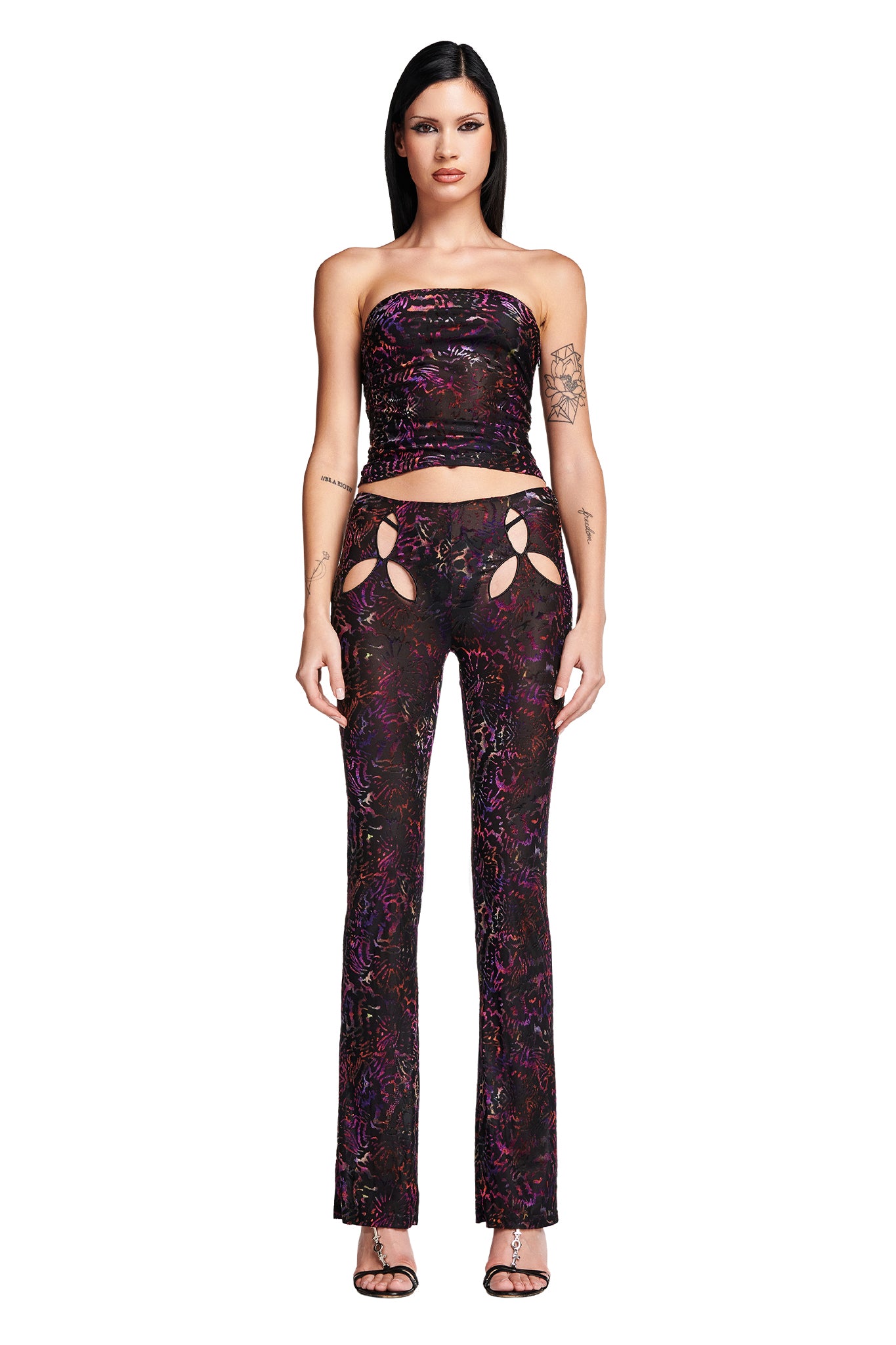 A woman with long black hair and tattoos on her arm stands facing forward, perfectly capturing the edgy style of I.AM.GIA. She is wearing a strapless black crop top and the I.AM.GIA AMINA PANT - ANIMAL BURN OUT, a pair of high-waisted, tight-fitting black pants featuring colorful patterns and cutout details on the thighs. Her outfit is completed with black high-heeled sandals.