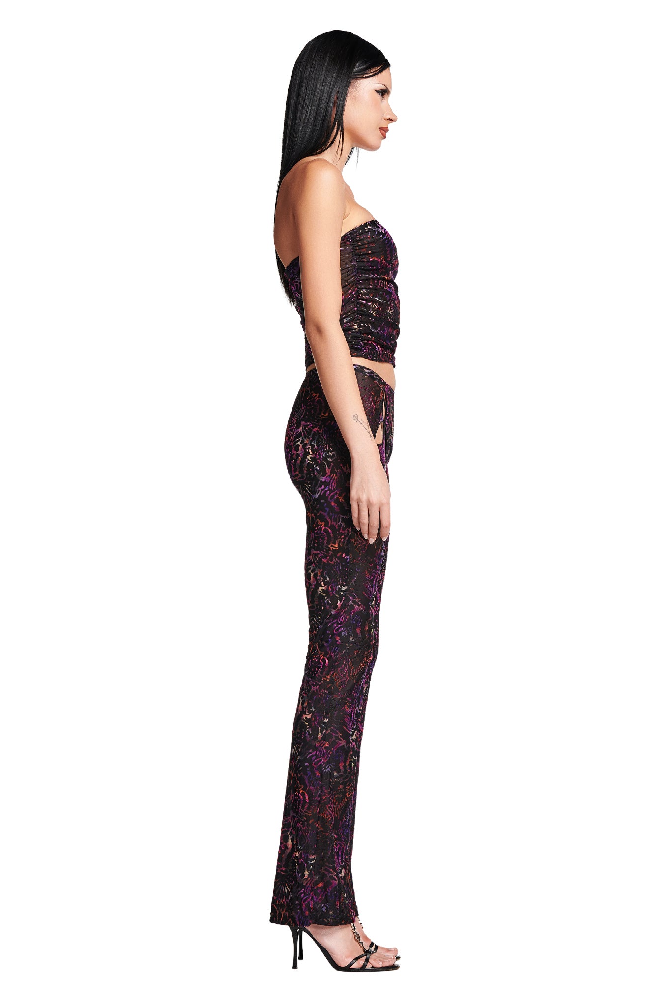 A woman with long dark hair stands in profile against a white background. She is wearing the AMINA PANT - ANIMAL BURN OUT from I.AM.GIA, featuring a dark, colorful design and paired with black high-heeled sandals. She appears to be looking slightly downward.