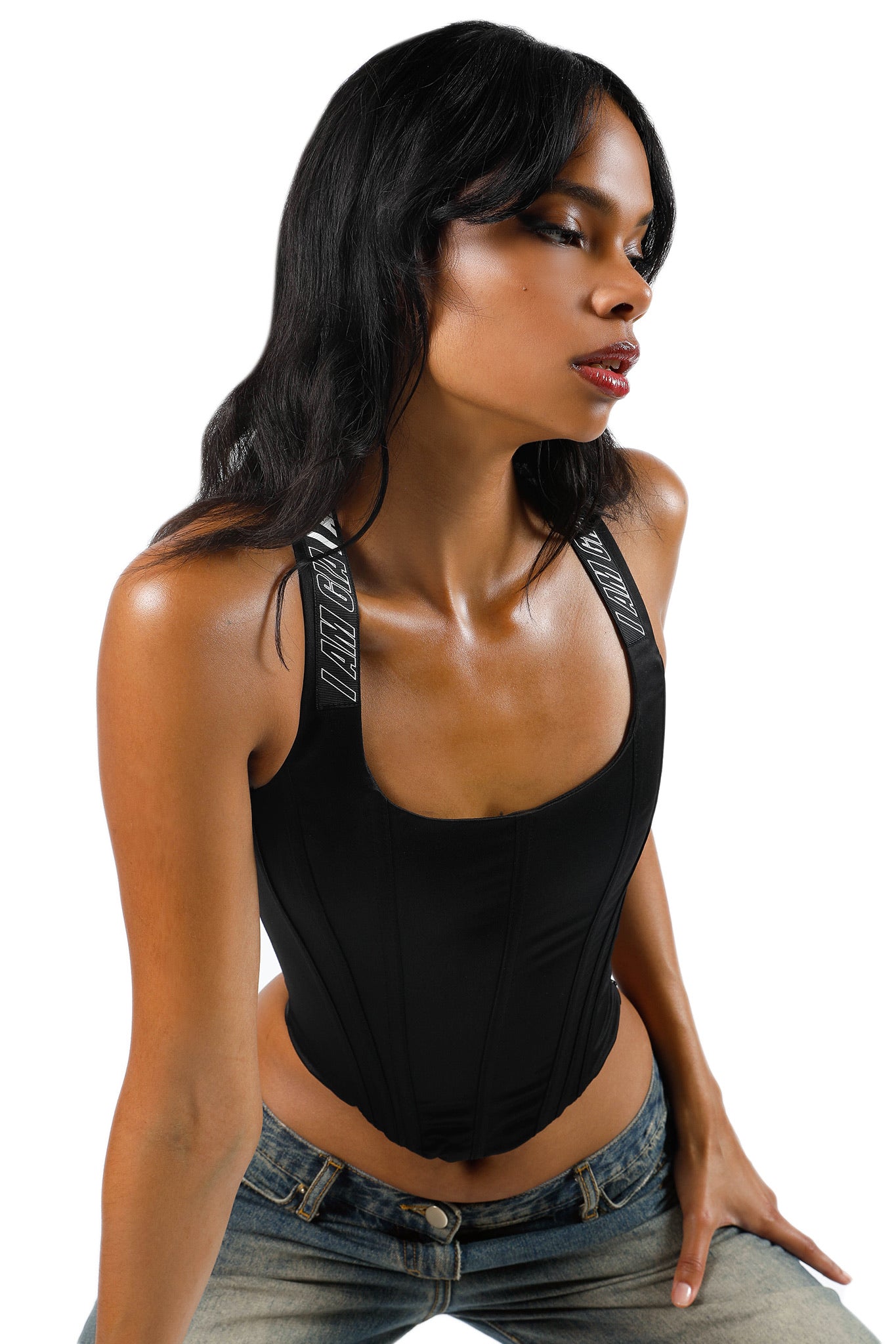 A woman with long dark hair dons the SIOBHAN CORSET - BLACK, a fitted top from I.AM.GIA featuring straps adorned with white text reading "I.AM.GIA." She pairs it stylishly with blue jeans. The image is set against a plain white background, and she gazes to the side with her lips slightly parted.