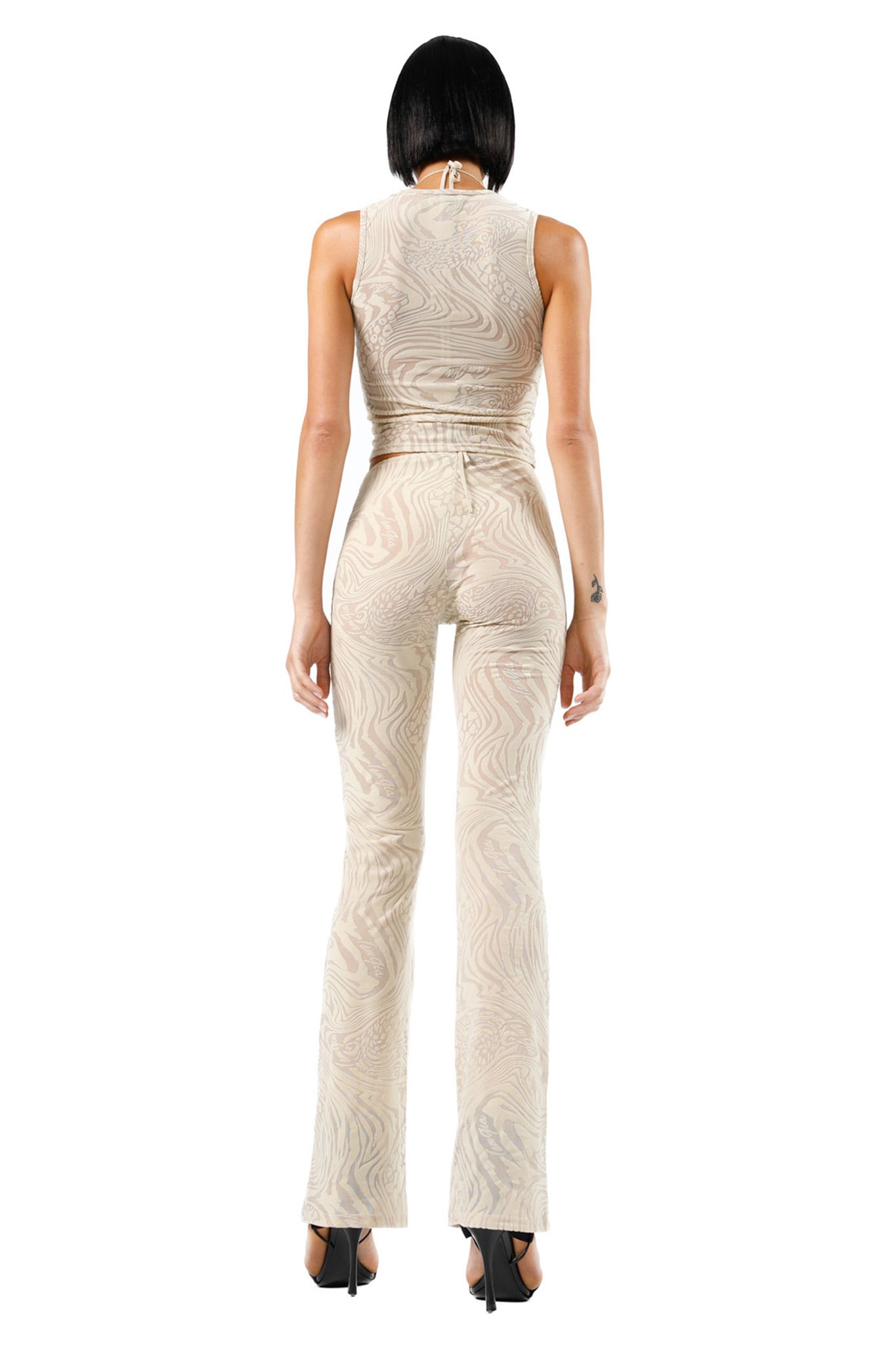 A person with short black hair is facing away from the camera, wearing a sleeveless, form-fitting beige outfit by I.AM.GIA featuring the AMINA PANT - TAN BURNOUT with a subtle wavy pattern. The ensemble includes high-waisted pants that flare at the bottom and black high heels, showcasing a tattoo on their right arm.