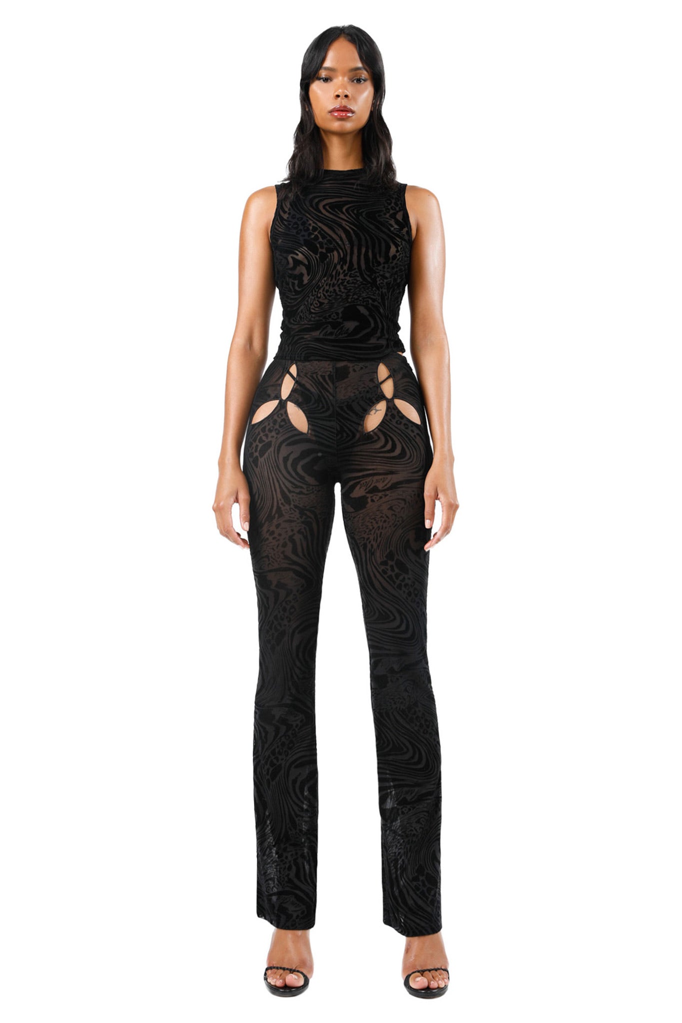 A woman stands facing forward wearing a sleeveless I.AM.GIA AMINA PANT - BLACK BURNOUT jumpsuit featuring cutouts on the hips. She has long dark hair and is wearing black open-toe heels. The background is plain white.
