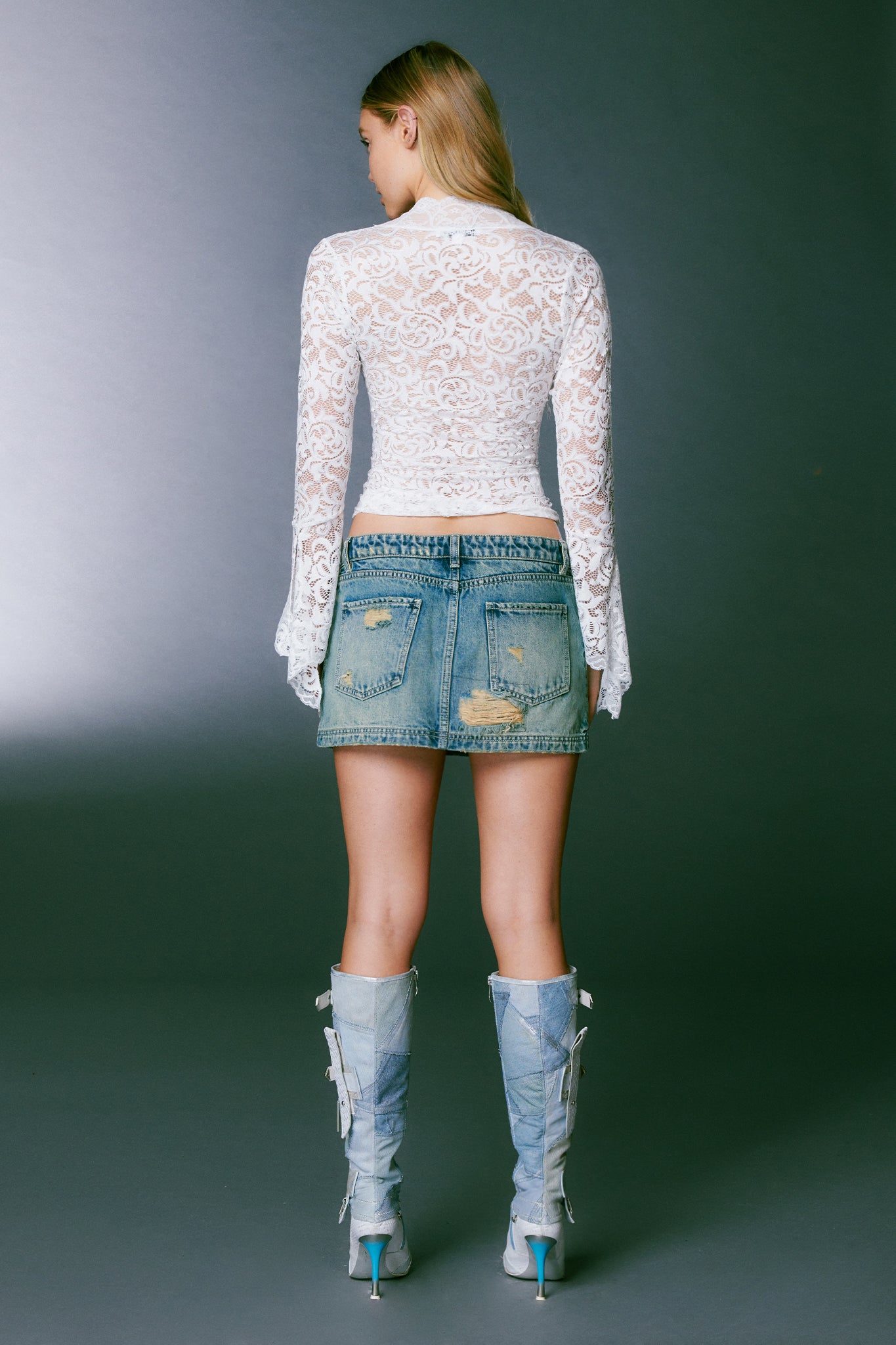 A person with long blonde hair stands facing away from the camera. They are wearing I.AM.GIA's GEMMA TOP - WHITE, a long-sleeved sheer lace top, paired with a distressed denim mini skirt and light blue high-heeled boots with white laces. The ensemble reflects I.AM.GIA's edgy fashion combinations. The background is a plain dark gray.
